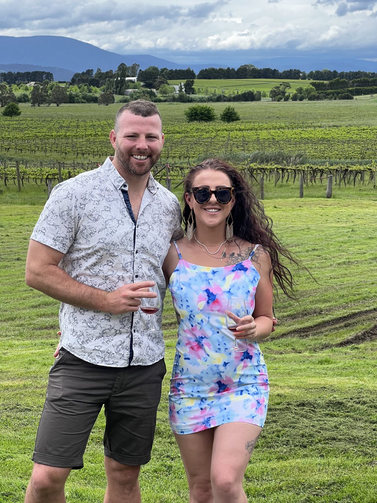 Dani and Alex with drinks in front of rows of grapevines